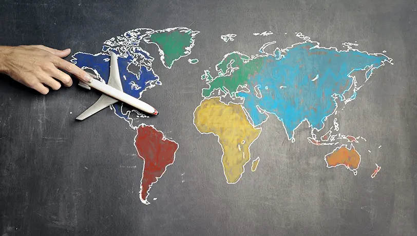 Starting a Business Overseas? A Quick Guide to Tax Considerations A male adult hand holds a scale model plane over a drawing of a colored world map on a chalkboard, illustrating a business overseas expansion.
