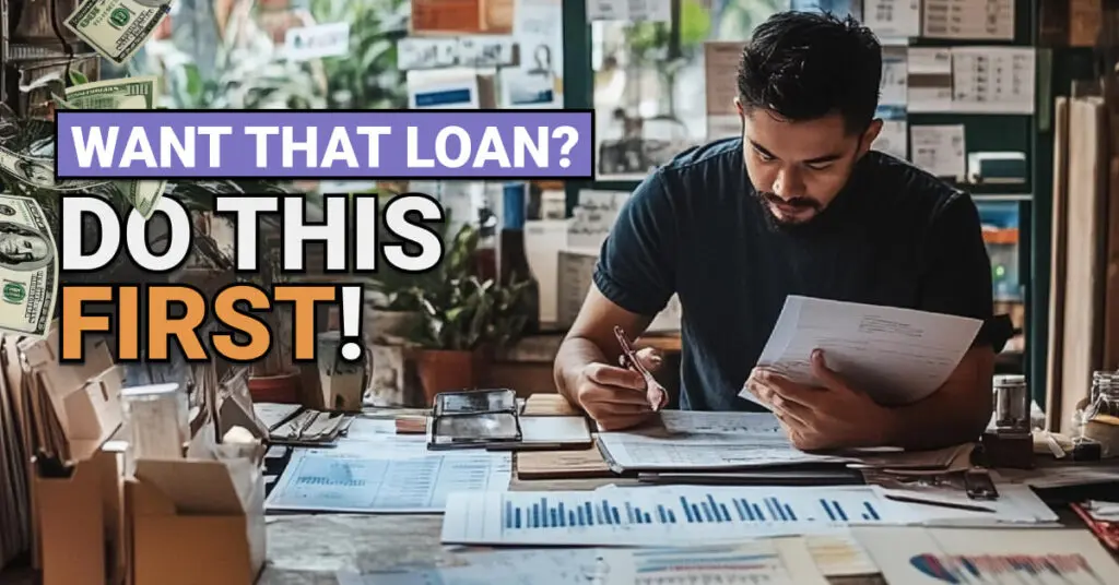 A focused man sits at a cluttered desk reviewing financial documents and charts in a home office or small business workspace. Text overlaid reads &ldquo;WANT THAT LOAN? DO THIS FIRST!&rdquo; with dollar bills floating around, suggesting financial preparation or planning for a loan.