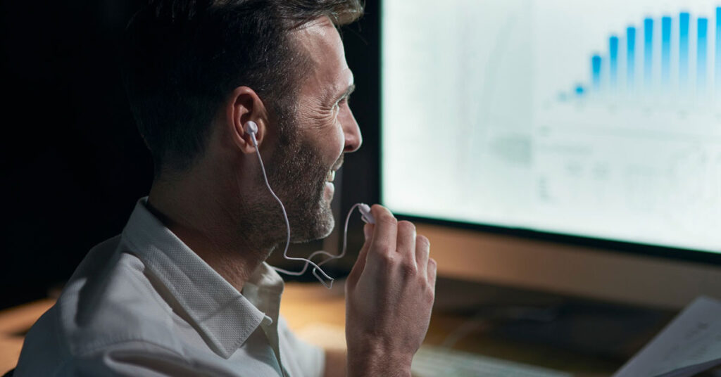 Smiling professional using earphones while analyzing financial charts on a computer, representing online investing and trading with broker guidance