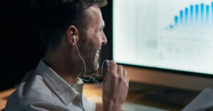 Smiling professional using earphones while analyzing financial charts on a computer, representing online investing and trading with broker guidance
