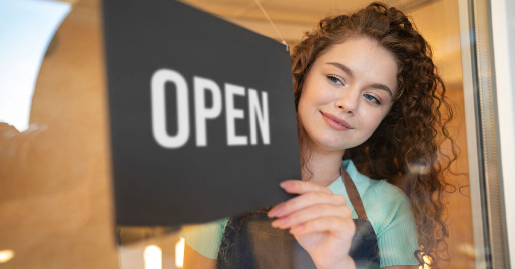 Young female entrepreneur smiling as she opens her new business, symbolizing the excitement and challenges of starting a new venture