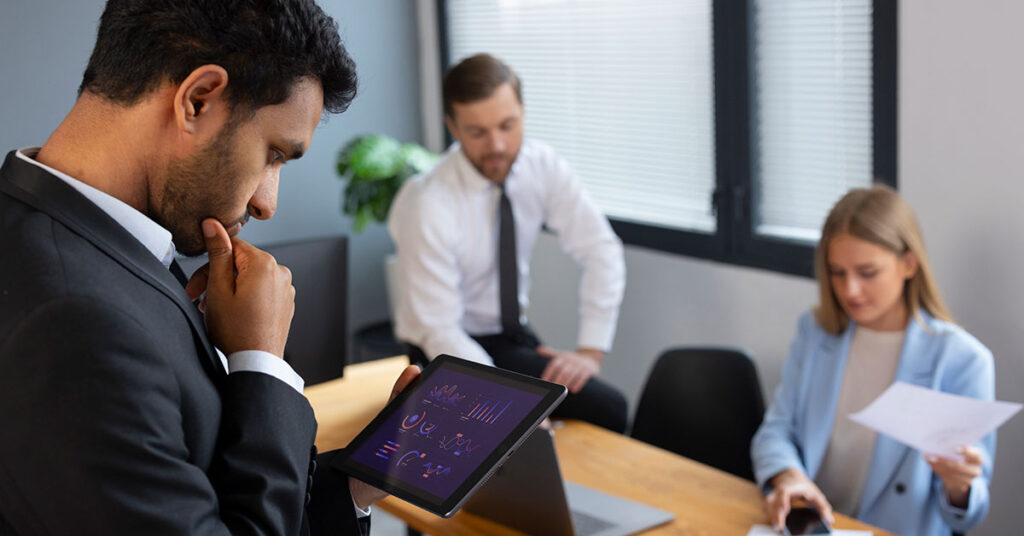 Business team in modern office, with a manager analyzing employee performance graphs on a digital tablet while colleagues review documents during a meeting.