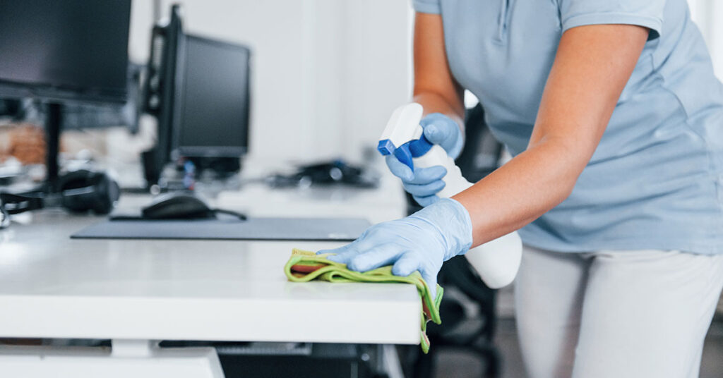 Person cleaning and disinfecting office desk with spray bottle and cloth while wearing protective gloves, ensuring a hygienic and sanitized commercial workspace.
