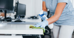 Person cleaning and disinfecting office desk with spray bottle and cloth while wearing protective gloves, ensuring a hygienic and sanitized commercial workspace.