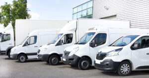 Row of white commercial vans and trucks parked outside a modern industrial building, showcasing a fleet of vehicles used for business and logistics operations.