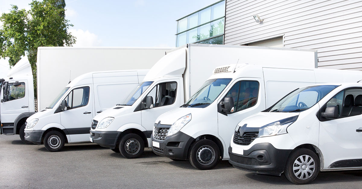 Row of white commercial vans and trucks parked outside a modern industrial building, showcasing a fleet of vehicles used for business and logistics operations.