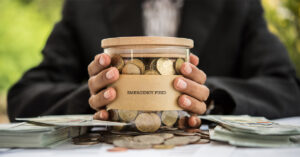 Person holding a glass jar labeled "Emergency Fund" filled with coins, surrounded by cash—symbolizing financial preparedness and accessing fast loans during emergencies.