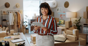 Smiling entrepreneur holding a tablet in a home office surrounded by packages and clothing—symbolizing the early stages of launching a small business.