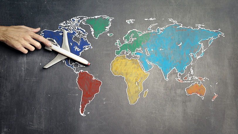A male adult hand holds a scale model plane over a drawing of a colored world map on a chalkboard, illustrating a business overseas expansion.