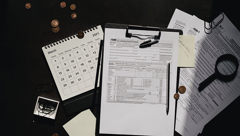 US Tax Forms rest among clipping board, a magnifying glass, a calendar, and coins.