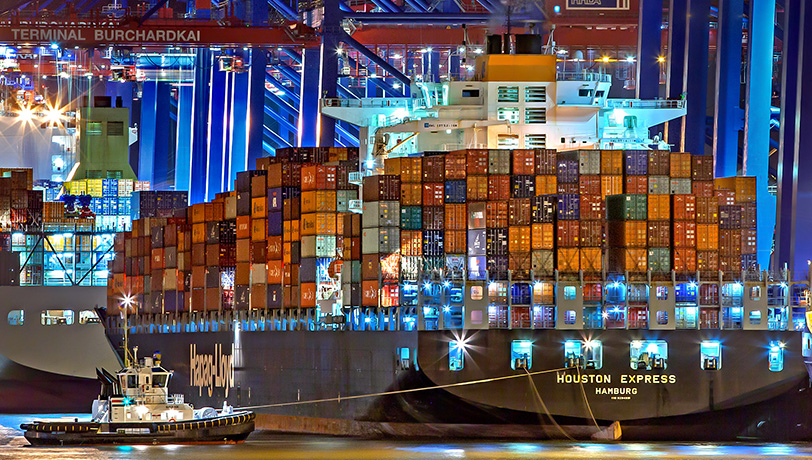 A cargo ship full of containers enters a docking port followed by a tugboat, depicting international trade, most of it led by overseas companies.