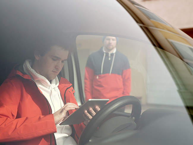 Windshield view of the driver of a small business commercial vehicle, accessing information with a tablet on his hands, while another employee appears standing in the background.