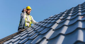 Construction worker in safety gear inspecting a metal roof under clear blue sky—representing roofing industry startups and funding opportunities.