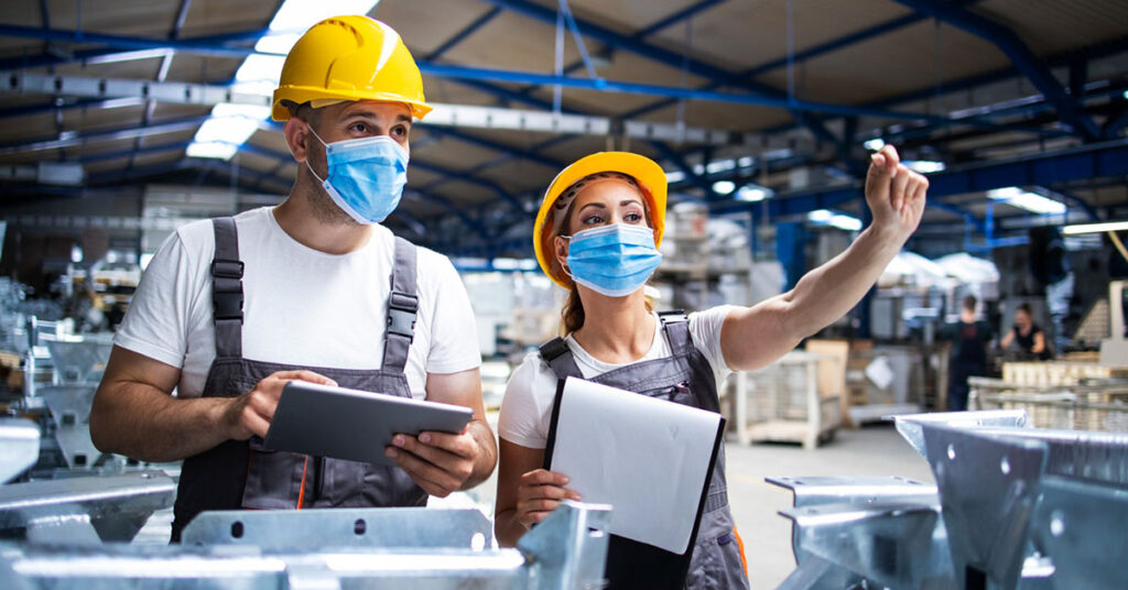 Two factory workers wearing safety helmets and face masks reviewing production plans on digital tablets inside an industrial manufacturing facility—representing industries reliant on working capital.