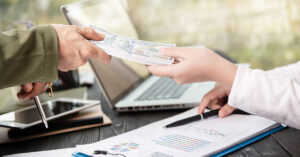Close-up of hands exchanging a stack of hundred-dollar bills over financial documents and a laptop, symbolizing quick cash loans and fast financing solutions.
