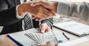 Close-up of a business handshake over loan documents and a stack of hundred-dollar bills, symbolizing the successful approval of a small business loan.