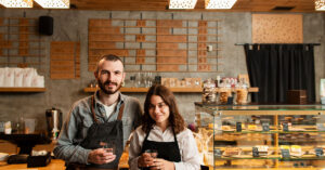 Smiling franchise business owners standing behind the counter of a modern café or bakery, representing the success and opportunities of franchise ownership and financing.