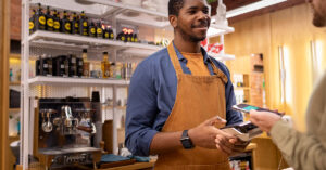 Smiling small business owner wearing an apron processing a contactless payment in a modern retail store, representing entrepreneurship and small business launch success.