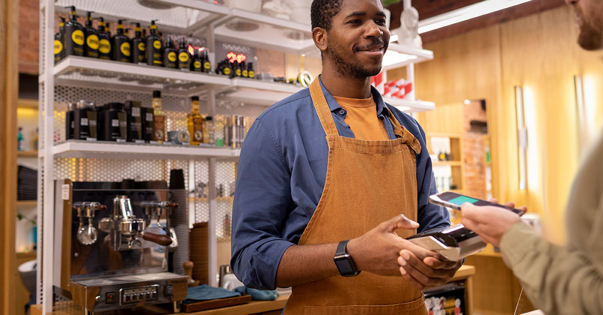 Smiling small business owner wearing an apron processing a contactless payment in a modern retail store, representing entrepreneurship and small business launch success.