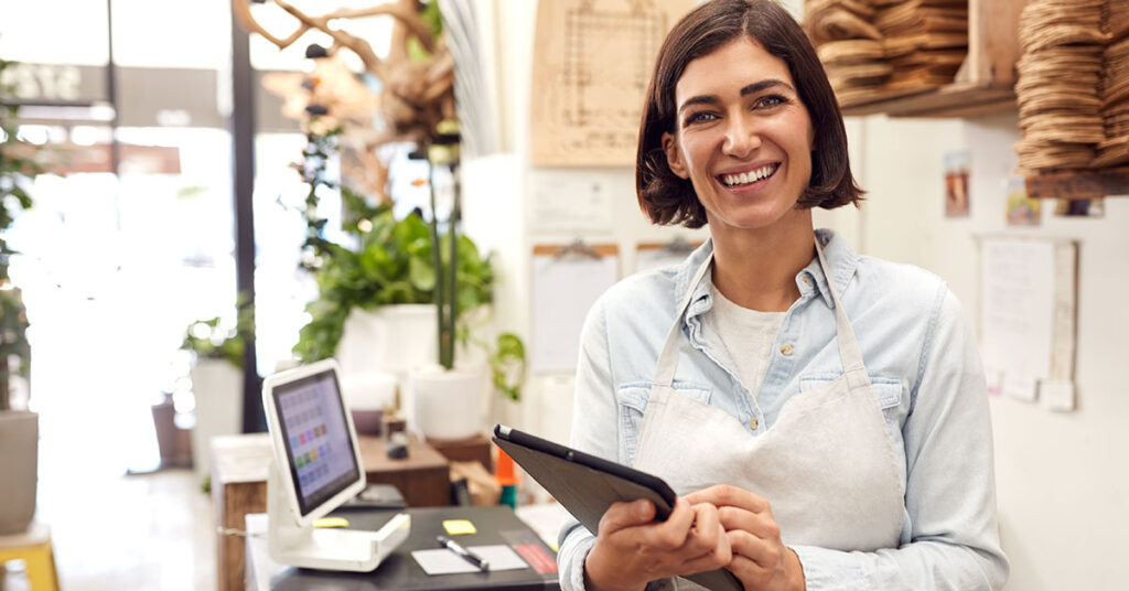 Smiling female entrepreneur standing behind the counter of a small business, holding a digital tablet, representing small business ownership and financing opportunities.