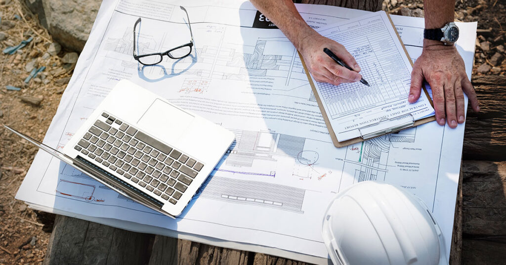 Construction professional filling out a contractor daily report on-site with architectural blueprints, laptop, and safety helmet, representing the financial planning needs of general contracting businesses.