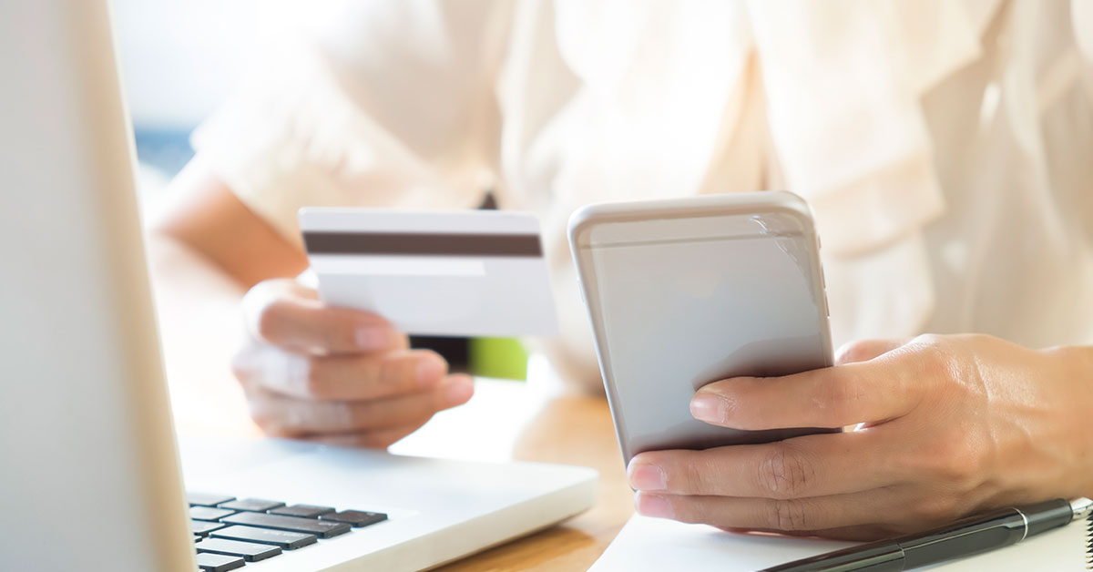 Close-up of a person holding a credit card and smartphone while using a laptop, representing online access to a business line of credit.