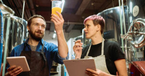 Two brewery workers examining a glass of craft beer while holding a tablet and clipboard inside a modern brewing facility, representing the craft beer industry and brewery business operations.