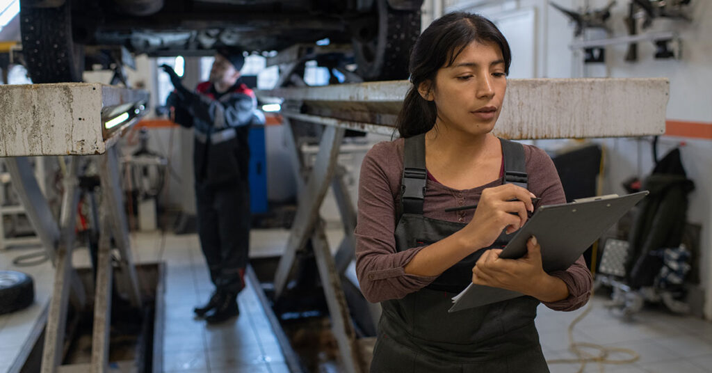 Female auto repair technician taking notes on a clipboard in a car service garage, representing the operational and financial needs of auto repair businesses.