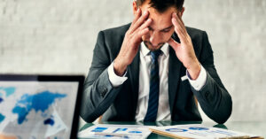 Frustrated businessman in a suit holding his head in his hands while reviewing financial charts, representing the stress of managing a small business with bad credit.